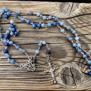 May include: A blue and silver rosary with a silver crucifix. The beads are made of blue agate and are graduated in size. The rosary is laid out on a wooden surface.