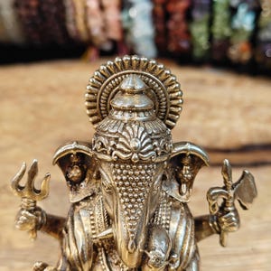 May include: A detailed, gold-colored statue of the Hindu deity Ganesha, holding a trident and an axe. The statue features intricate detailing on the crown, trunk, and jewelry. The background is blurred, with colorful beads.