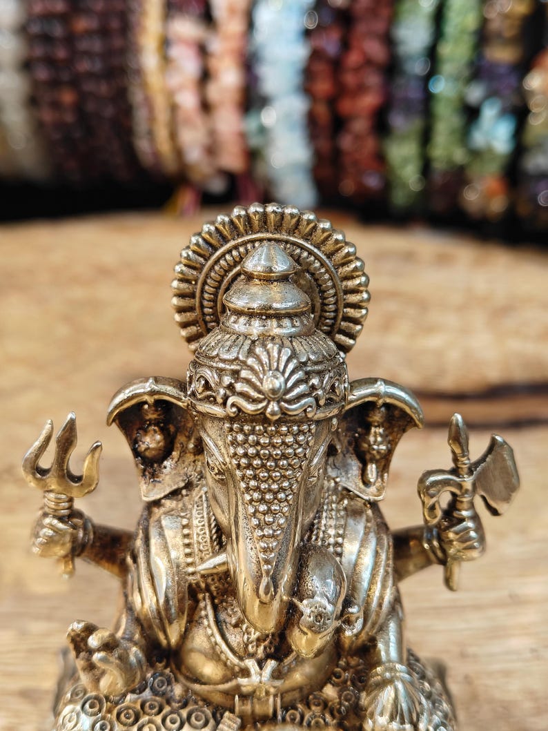 May include: A detailed, gold-colored statue of the Hindu deity Ganesha, holding a trident and an axe. The statue features intricate detailing on the crown, trunk, and jewelry. The background is blurred, with colorful beads.