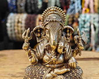 Puede incluir: Una detallada estatua de latón de la deidad hindú Ganesha, sentado en una postura meditativa. La estatua presenta detalles intrincados, incluyendo una corona, múltiples brazos sosteniendo varios objetos y un ratón en la base. El fondo muestra una exhibición de pulseras de cuentas.