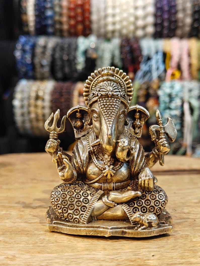 May include: A detailed brass statue of the Hindu deity Ganesha, seated in a meditative pose. The statue features intricate detailing, including a crown, multiple arms holding various objects, and a mouse at the base. The background shows a display of beaded bracelets.