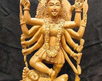 Può includere: Scultura in legno dettagliata di una divinità con molte braccia, probabilmente Kali, con intagli intricati. La figura è adornata da una corona e una collana di volti, in piedi su un'altra figura. La scultura è in legno chiaro.