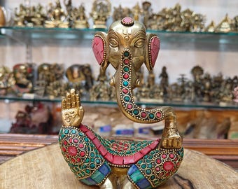 May include: A brass Ganesh statue with a detailed design. The elephant-headed deity is adorned with red, green, and blue accents. The statue is displayed on a wooden surface, with other religious items visible in the background.
