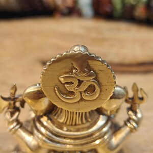 May include: A detailed brass statue of a deity, featuring a prominent circular emblem with the Om symbol. The statue has multiple arms and is adorned with intricate details. The overall color is a warm gold, with a focus on religious and spiritual themes.