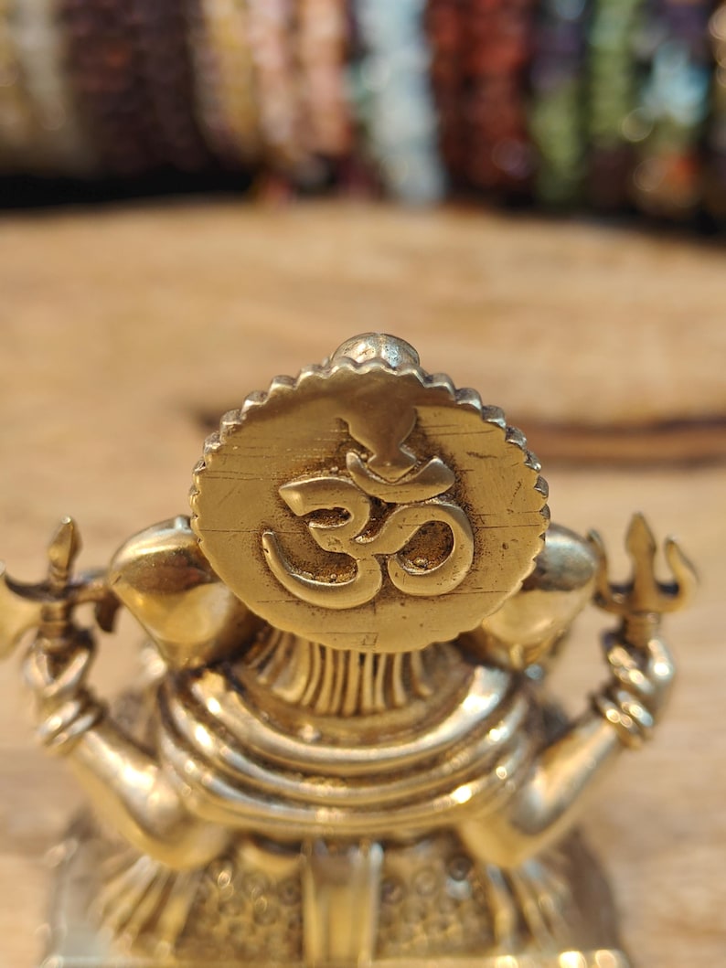 May include: A detailed brass statue of a deity, featuring a prominent circular emblem with the Om symbol. The statue has multiple arms and is adorned with intricate details. The overall color is a warm gold, with a focus on religious and spiritual themes.
