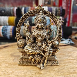 Puede incluir: Una detallada estatua de latón de una deidad sentada, con intrincadas tallas y detalles ornamentados. La figura está adornada con una corona y joyas, sobre un fondo arqueado. La estatua se exhibe sobre una superficie de madera.