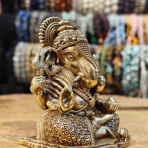 May include: A detailed, golden-colored statue of the Hindu deity Ganesha, seated in a meditative pose. The statue features intricate carvings, a large elephant head, and multiple arms holding symbolic objects. The background is blurred, with various beaded bracelets.