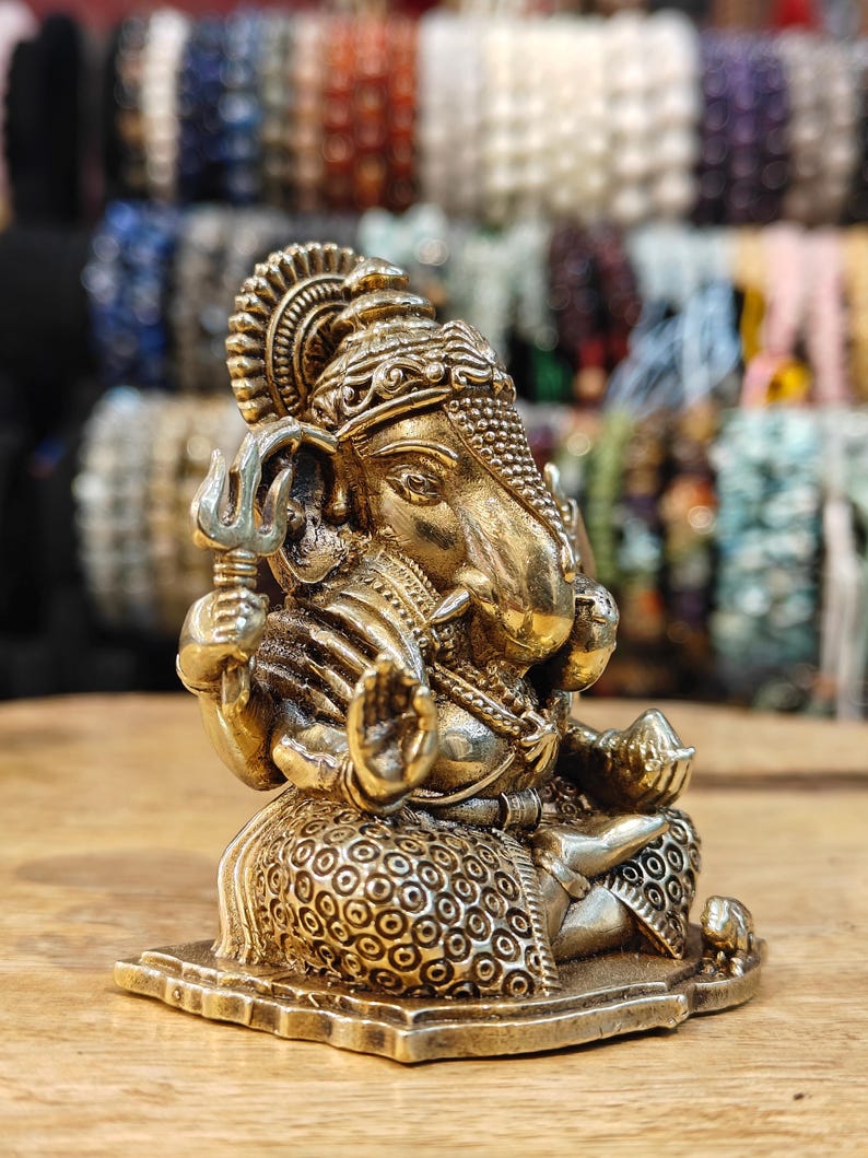 May include: A detailed, golden-colored statue of the Hindu deity Ganesha, seated in a meditative pose. The statue features intricate carvings, a large elephant head, and multiple arms holding symbolic objects. The background is blurred, with various beaded bracelets.