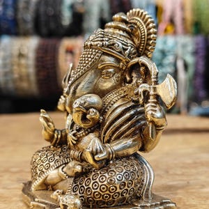May include: A detailed brass statue of the Hindu deity Ganesha, seated with multiple arms, holding various symbolic objects. The statue has intricate detailing, including a crown and ornate clothing. The background is blurred, with a wooden surface in the foreground.