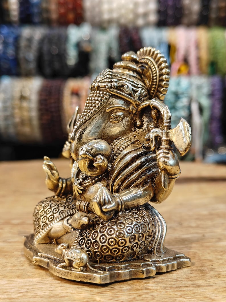 May include: A detailed brass statue of the Hindu deity Ganesha, seated with multiple arms, holding various symbolic objects. The statue has intricate detailing, including a crown and ornate clothing. The background is blurred, with a wooden surface in the foreground.