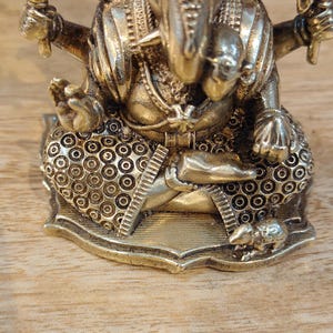 May include: A detailed brass statue of the Hindu deity Ganesha, seated in a meditative pose. The statue features intricate detailing, including a crown, jewelry, and a trident. The base is decorated with circular patterns. The statue is on a wooden surface.