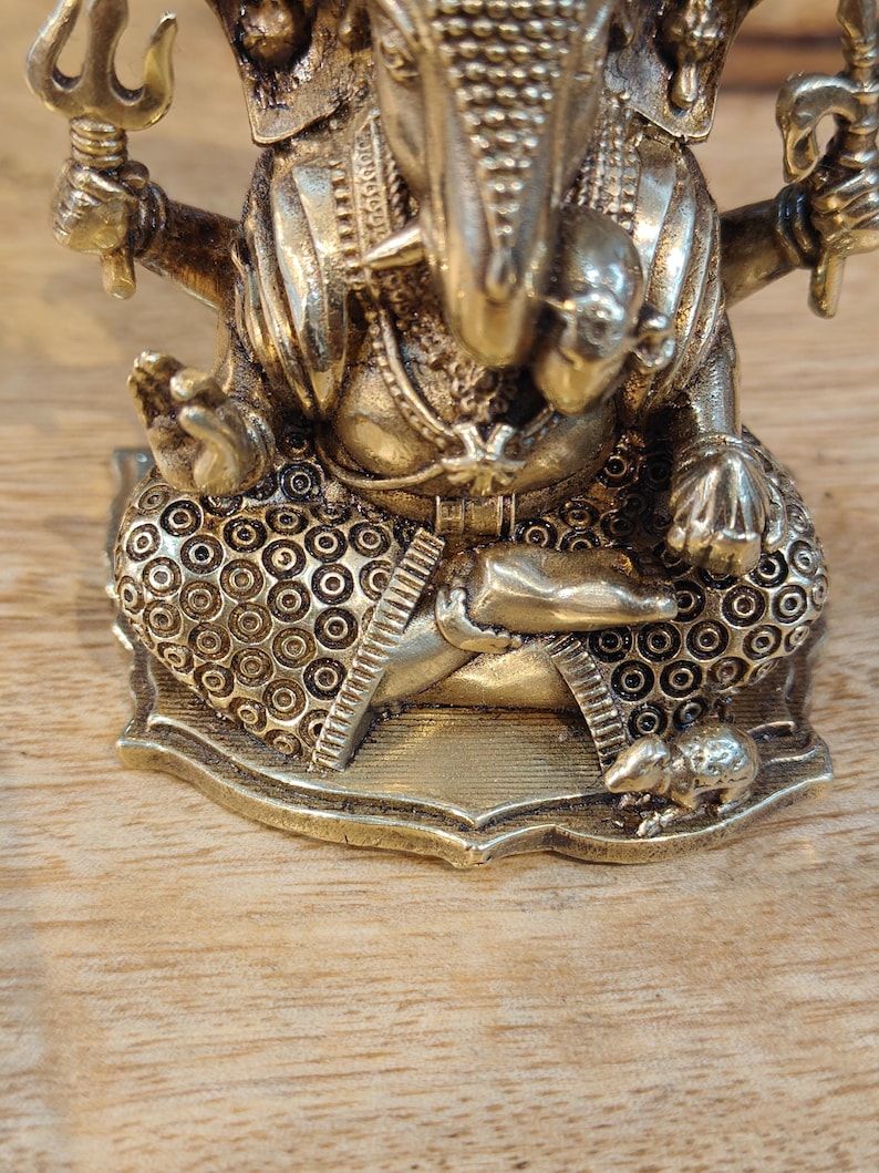 May include: A detailed brass statue of the Hindu deity Ganesha, seated in a meditative pose. The statue features intricate detailing, including a crown, jewelry, and a trident. The base is decorated with circular patterns. The statue is on a wooden surface.