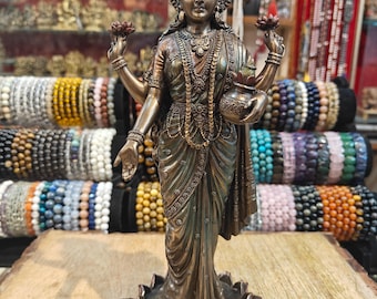 May include: Bronze-colored statue of a multi-armed deity, adorned with intricate jewelry and a headdress. The figure holds lotus flowers and a pot, standing on a lotus flower base. A small bowl with decorative elements sits at the base. The background features shelves of bracelets.