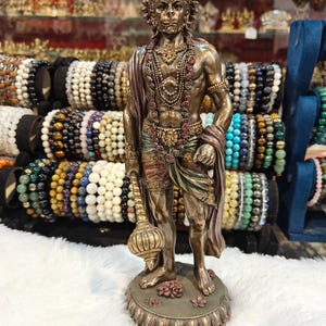 Puede incluir: Estatua de bronce de una deidad, adornada con intrincadas joyas y una corona. La figura sostiene una maza y está envuelta en un chal. La estatua se alza sobre una base decorativa con detalles florales. Una colección de pulseras de cuentas es visible en el fondo.