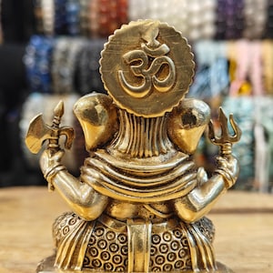 May include: A brass statue of a deity with multiple arms, holding various objects. The statue features a large circular emblem with the Om symbol. The figure is detailed with intricate carvings and is set on a square base.