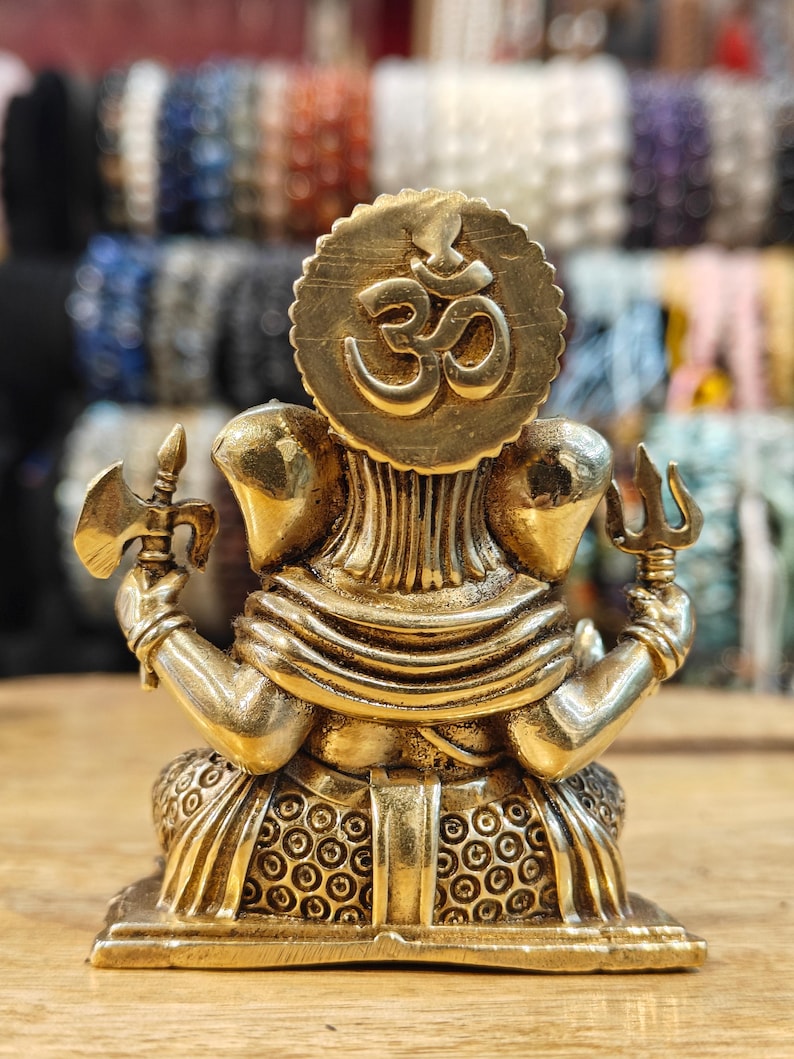 May include: A brass statue of a deity with multiple arms, holding various objects. The statue features a large circular emblem with the Om symbol. The figure is detailed with intricate carvings and is set on a square base.