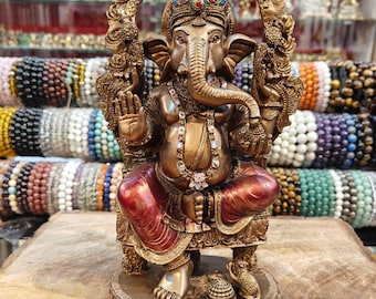 Puede incluir: Una estatua de color bronce del dios hindú Ganesha, sentado en un trono. La estatua presenta detalles intrincados, incluyendo una corona, joyas y una gran cabeza de elefante. Las túnicas de la deidad son rojas. La estatua se exhibe sobre una superficie de madera.