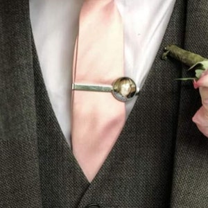 May include: A light pink tie with a silver tie clip featuring a round photo. The tie is worn with a white shirt and a dark grey suit.