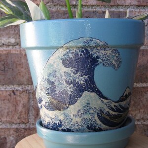May include: A blue ceramic planter with a graphic of a wave, painted in shades of blue and white. The planter is sitting on a wooden stand with three legs.