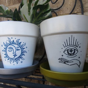 May include: Two white ceramic flower pots with blue and black designs. One pot has a sun and moon design, the other has an eye with a star inside and a hand reaching out. The pots are on a black metal stand.