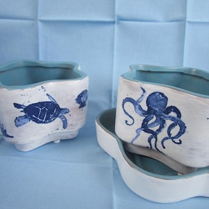 May include: White ceramic planters with a blue rim and sea creature designs. One planter features a blue turtle, starfish, and seashell, while another showcases a blue octopus. The planters are on a light blue surface.