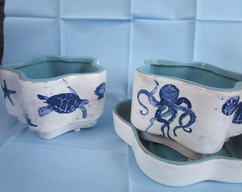 Può includere: Fioriere in ceramica bianca con bordo blu e disegni di creature marine. Una fioriera presenta una tartaruga blu, stelle marine e conchiglie, mentre un'altra mostra un polpo blu. Le fioriere sono su una superficie azzurra.