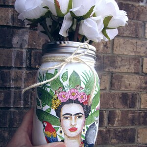 Pu&ograve; includere: Un barattolo di vetro bianco con un disegno dipinto di Frida Kahlo, foglie tropicali e un pappagallo. Il barattolo &egrave; decorato con fiori bianchi e uno spago.