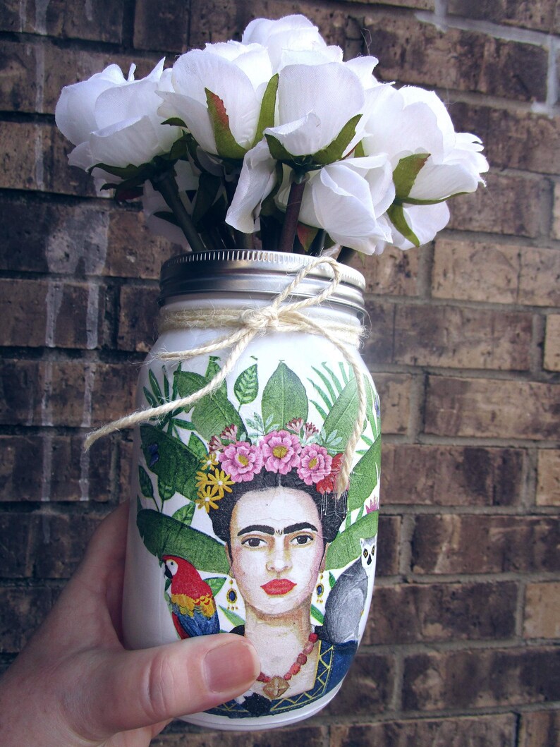 Pu&ograve; includere: Un barattolo di vetro bianco con un disegno dipinto di Frida Kahlo, foglie tropicali e un pappagallo. Il barattolo &egrave; decorato con fiori bianchi e uno spago.