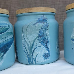 May include: Three blue glass jars with wooden lids, each decorated with a different sea creature. The first jar features a whale, the second a seahorse, and the third a sea turtle.