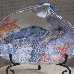 May include: A decorative seashell with a painted sea turtle design. The turtle is blue and white, surrounded by coral and aquatic plants. The shell has a silver rim and is displayed on a black metal stand.