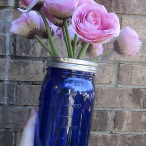 May include: A cobalt blue mason jar vase filled with pink flowers. The jar has measurement markings in milliliters and a silver lid. The flowers are a light pink color with green stems. The background is a brick wall.
