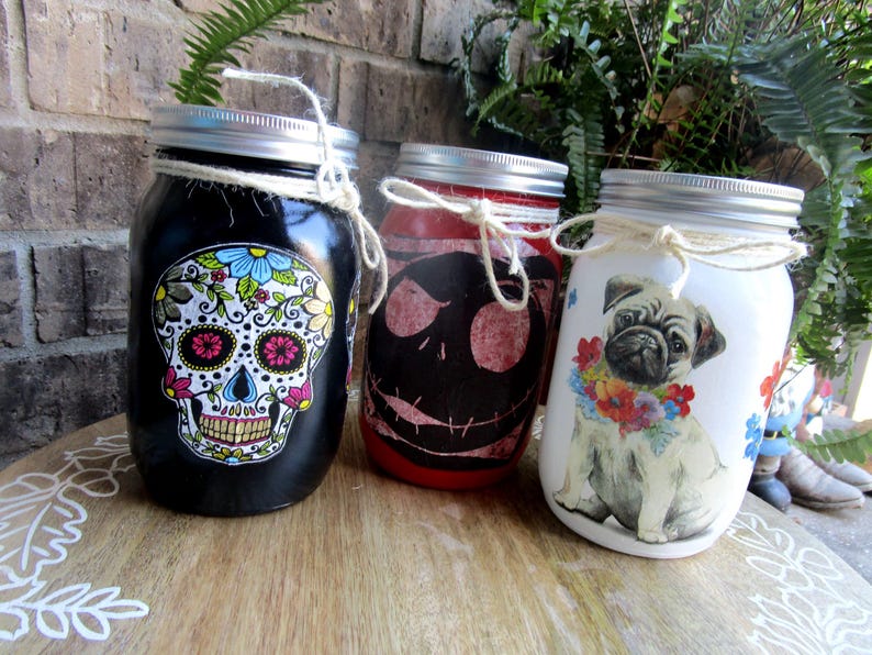Pu&ograve; includere: Tre barattoli di vetro decorati con diversi disegni. Il primo barattolo &egrave; nero con un disegno di teschio di zucchero colorato. Il secondo barattolo &egrave; rosso con un disegno di Jack Skellington in bianco e nero. Il terzo barattolo &egrave; bianco con un carlino che indossa una ghirlanda di fiori.