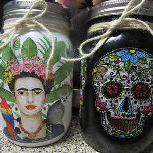Pu&ograve; includere: Due barattoli di vetro decorati con immagini colorate. Il barattolo a sinistra presenta un ritratto di Frida Kahlo con piante tropicali e un pappagallo. Il barattolo a destra presenta un teschio di zucchero bianco e nero con dettagli floreali.