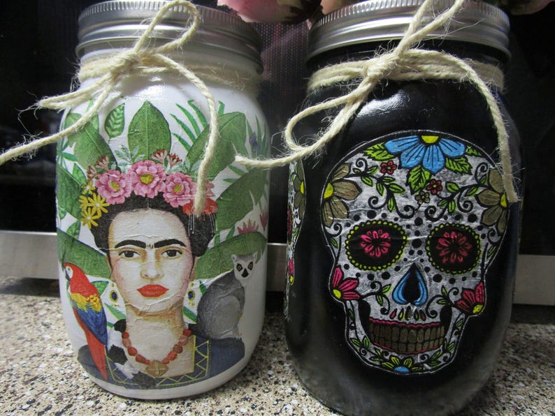 Pu&ograve; includere: Due barattoli di vetro decorati con immagini colorate. Il barattolo a sinistra presenta un ritratto di Frida Kahlo con piante tropicali e un pappagallo. Il barattolo a destra presenta un teschio di zucchero bianco e nero con dettagli floreali.