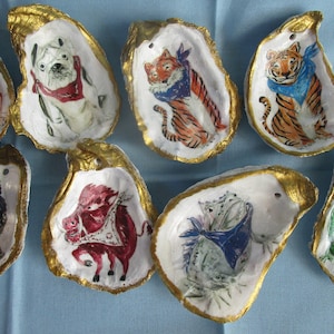 May include: A collection of hand-painted oyster shells with gold rims. Each shell features a different animal illustration, including a bulldog, pug, tiger, wolf, bull, iguana, and alligator. The artwork is colorful and detailed.