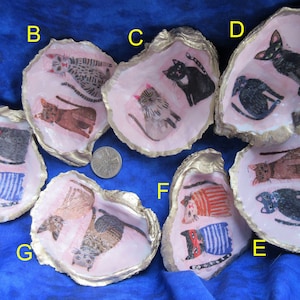 May include: Eight oyster shells with gold trim, each painted with a different cat design. The cats are painted in various colours, including black, white, brown, and grey. The shells are arranged in a circle on a blue fabric background.