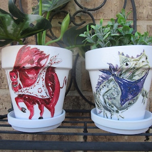 Peut inclure: Deux pots de fleurs en céramique blanche avec des motifs peints à la main. Un pot représente un sanglier rouge portant un bandana blanc, l'autre pot représente un lézard vert portant un bandana violet.