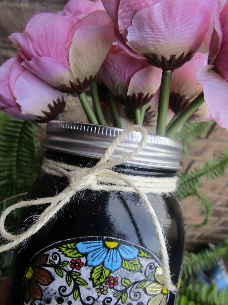 Pu&ograve; includere: Un barattolo di vetro nero con un designo decorativo a forma di teschio dipinto sul davanti. Il barattolo &egrave; legato con uno spago e riempito con fiori di seta rosa.