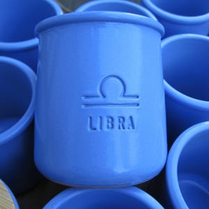 Puede incluir: Un juego de seis tazas de cerámica azul con signos del zodíaco. La taza del centro presenta el símbolo del zodíaco de Libra y la palabra "LIBRA" en relieve en la parte delantera.