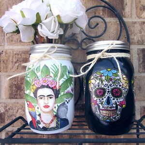 Pu&ograve; includere: Due barattoli di vetro decorati con disegni dipinti. Il barattolo a sinistra presenta un ritratto di Frida Kahlo con piante tropicali e un pappagallo. Il barattolo a destra &egrave; nero con un disegno di teschio di zucchero del Giorno dei Morti.