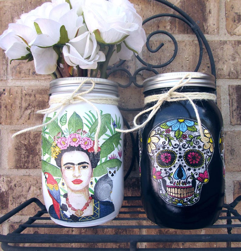 Pu&ograve; includere: Due barattoli di vetro decorati con disegni dipinti. Il barattolo a sinistra presenta un ritratto di Frida Kahlo con piante tropicali e un pappagallo. Il barattolo a destra &egrave; nero con un disegno di teschio di zucchero del Giorno dei Morti.