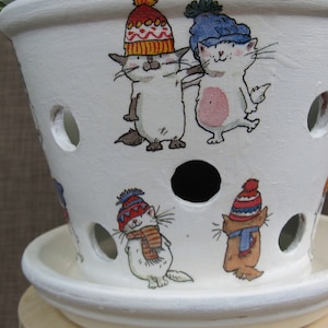 Puede incluir: Maceta de cerámica blanca con agujeros de drenaje, con un diseño caprichoso de gatos con gorros de invierno y bufandas. La maceta tiene un platillo a juego.