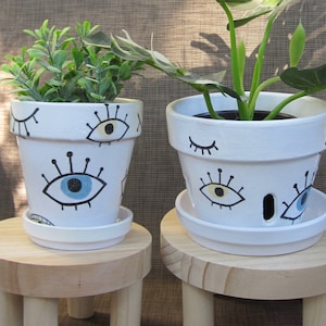 May include: Two white ceramic plant pots with eye designs. The pots are on wooden stands. One pot has a blue eye and the other has a yellow eye. The plants are a green leafy plant and a monstera plant.