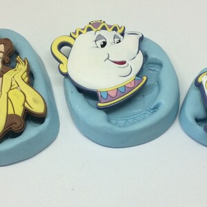 Peut inclure: Trois moules en silicone pour la fabrication de décorations de gâteaux. Les moules sont bleus et présentent des personnages du film Disney La Belle et la Bête. Les personnages sont Belle, Mme Samovar et Chip.