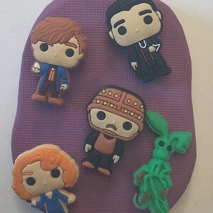 May include: Five colorful, cartoon-style figurines on a purple background. The figurines include a brown-haired boy holding a white egg, a man with black hair wearing a black suit and a red tie, a green creature with long legs, a woman with orange hair wearing a blue dress, and a brown and yellow robot-like figure.