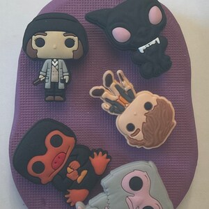 May include: A set of six colorful character charms, including a black cat, a brown-haired figure, a gray creature, a brown-haired figure with a wand, and a brown and orange figure. The charms are on a purple background.