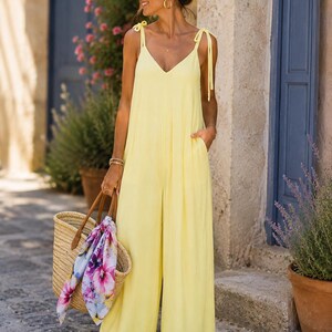 Peut inclure: Une combinaison sans manches jaune pâle avec un décolleté en V, des bretelles à nouer et un pantalon large avec des poches latérales. Un sac fourre-tout en paille tressée avec des anses en cuir marron et un foulard à motif floral coloré.