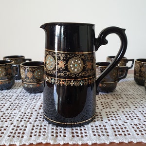 May include: A black ceramic pitcher and cups with gold and colorful accents. The pitcher and cups have a decorative design with gold trim and colorful jewels.