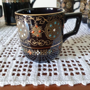 Pode incluir: Uma chávena de chá preta e dourada com um desenho floral e detalhes brancos. A chávena está sobre um tapete de crochê branco.