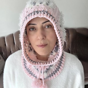 May include: A pink and gray crocheted hood with a matching cowl and pom-poms. The hood features a fluffy white trim and a drawstring closure. The cowl has a striped pattern in pink, gray, and white.